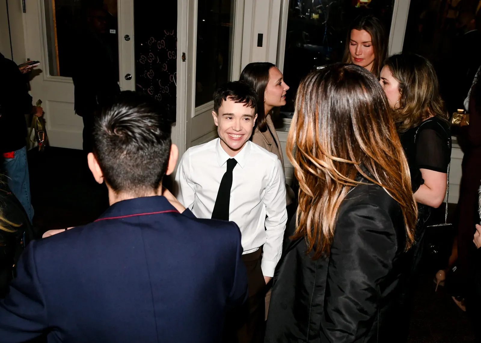 Elliot Page, dressed smartly in a white shirt and black tie, is smiling and engaging with guests at the Gucci New York Wooster Boutique reopening event.