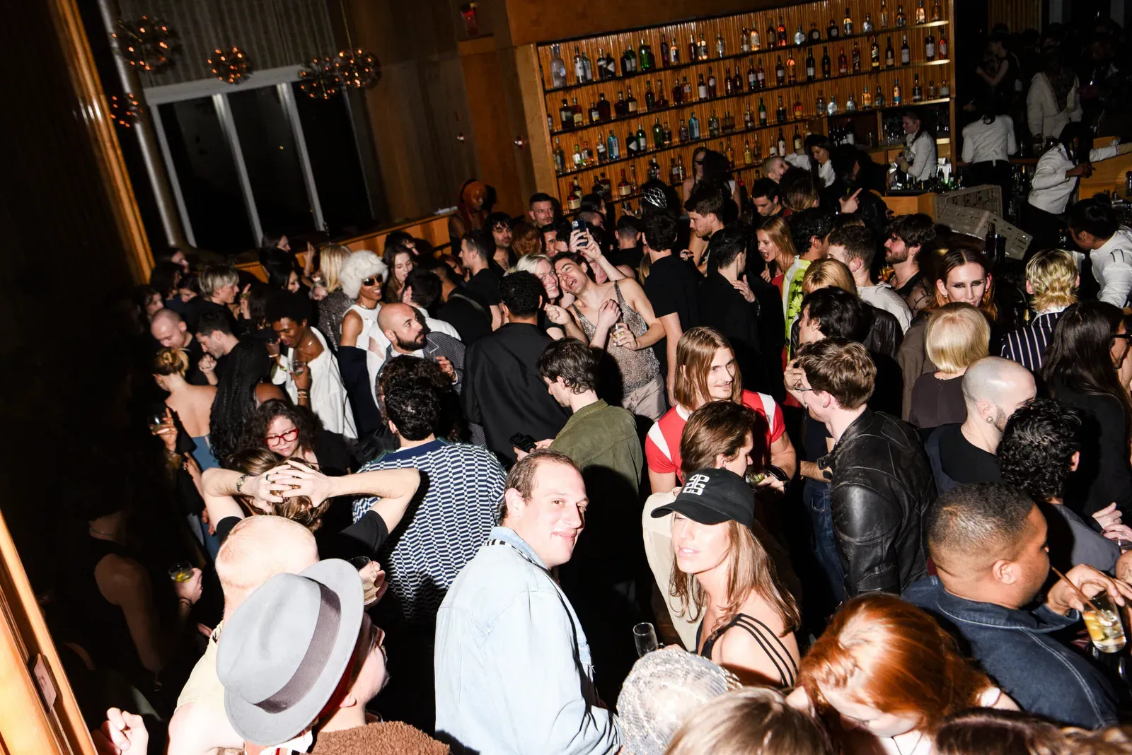 A bustling crowd at the NYFW afterparty for Ludovic de Saint Sernin. Guests are mingling and enjoying drinks, with a diverse array of fashion styles on display, set against a backdrop of a well-stocked bar with amber lighting.