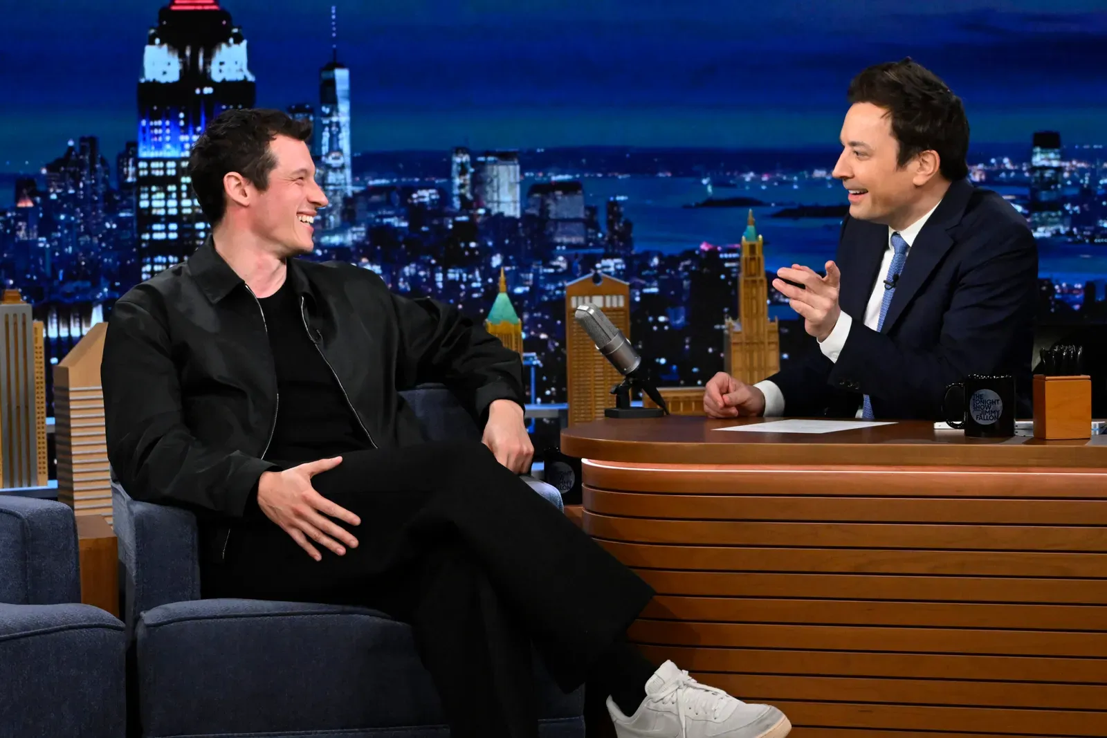 Actor Callum Turner and Jimmy Fallon engaged in a lively conversation on a talk show set, with one man seated across from the host, both laughing and gesturing.