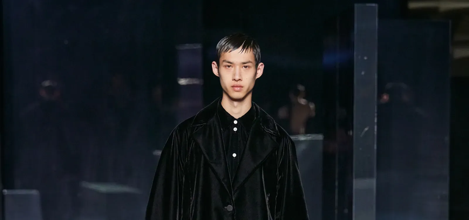 A male model walks down the runway in EENK's AW24 collection, wearing a shiny black shirt jacket over a white shirt, embodying the brand's minimalist and elegant design philosophy against the reflective runway environment.
