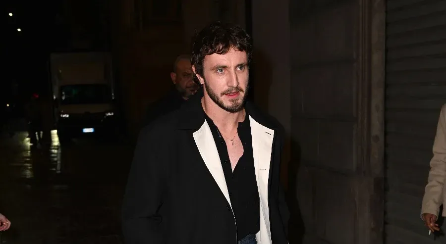 Paul Mescal walking confidently in a stylish outfit featuring a black coat over a white shirt, paired with classic jeans and black loafers with white socks.