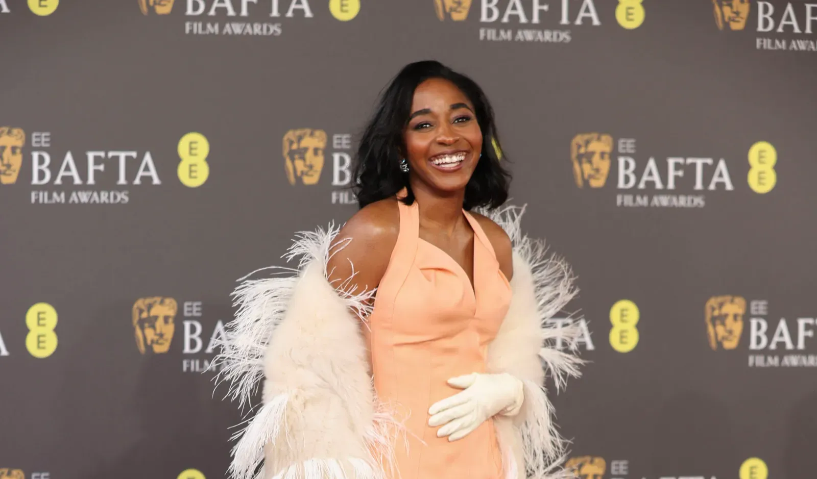 Ayo Edebiri stands on the red carpet at the 2024 BAFTA Film Awards, radiant in a bespoke Bottega Veneta peach gown with a luxuriously plumed shawl.
