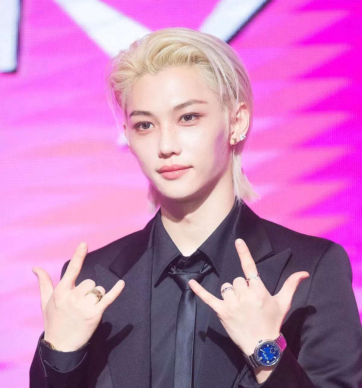 Felix of Stray Kids at a press conference, dressed in a Louis Vuitton black tuxedo and accessorized with fine jewelry, against a pink backdrop.