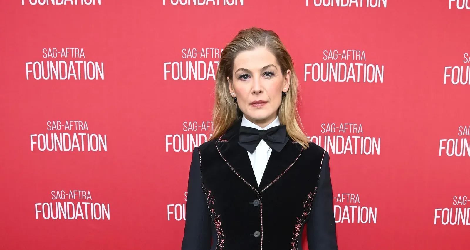 Actress Rosamund Pike at the SAG-AFTRA Foundation event in Los Angeles, wearing a black velvet dress with pink embroidery, a white high-collar shirt, and a black bow tie.