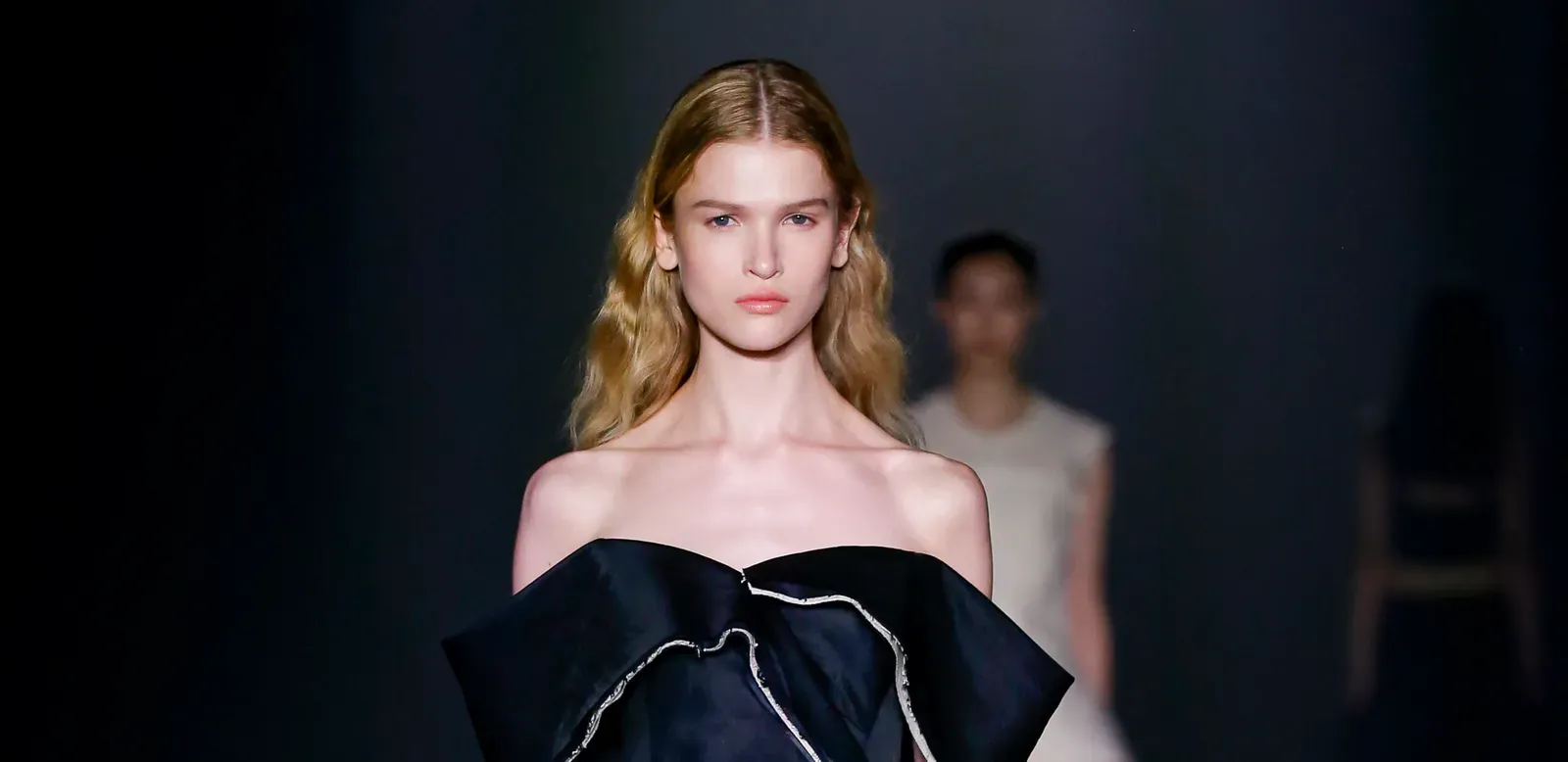A model elegantly parades down the runway in a Jason Wu FW24 off-the-shoulder gown with a dramatic, oversized ruffled neckline, edged with a distinctive white trim, showcasing a blend of classic femininity and modern design.