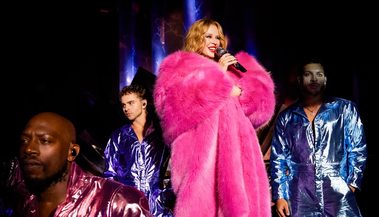 Kylie Minogue on stage holding a microphone, wearing a vibrant pink fur coat, with backup dancers in metallic outfits in the background.