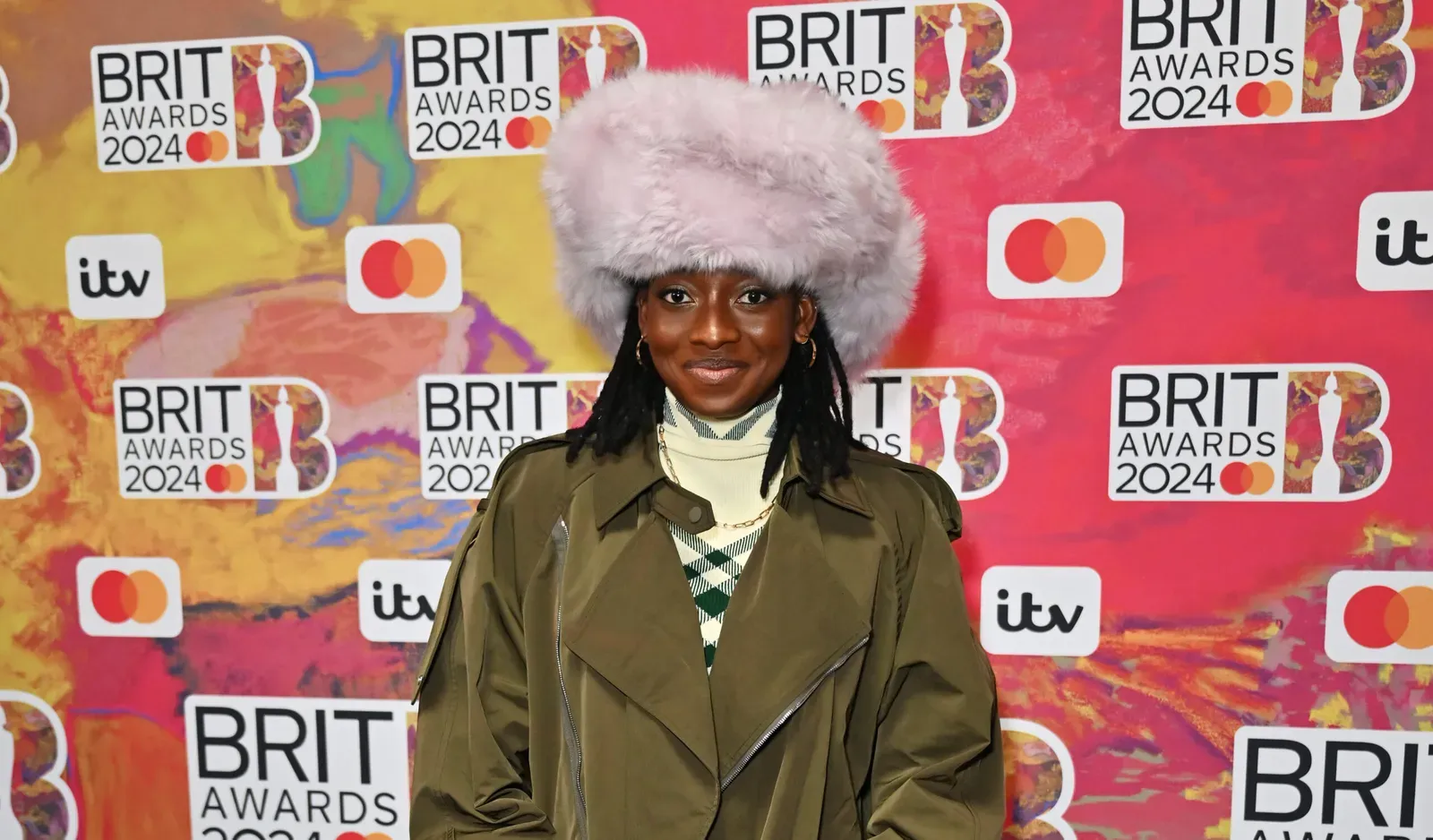 Little Simz strikes a pose at the BRIT Awards 2024, wearing a Burberry wool trench coat, ivy suede trousers, and accessorized with a shearling hat and leather gloves.