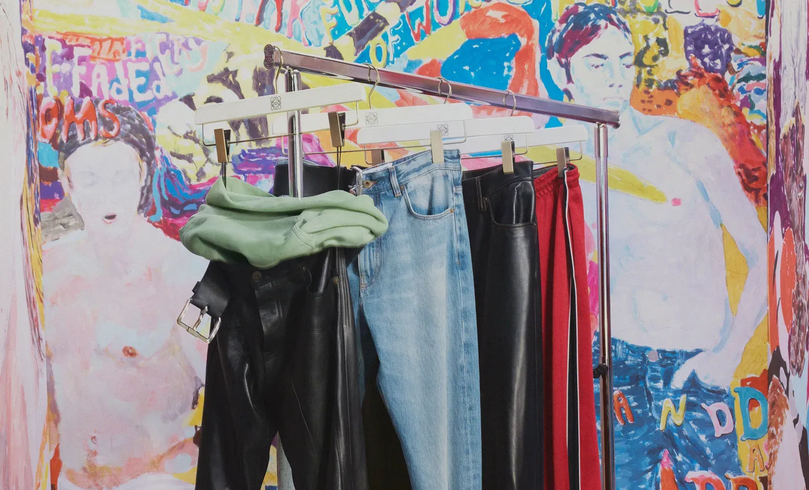 A selection of LOEWE's Fall/Winter 2024 menswear collection hangs against a backdrop of colorful, expressive artwork, featuring a variety of pants including green leather, classic denim, and bold red stripes.