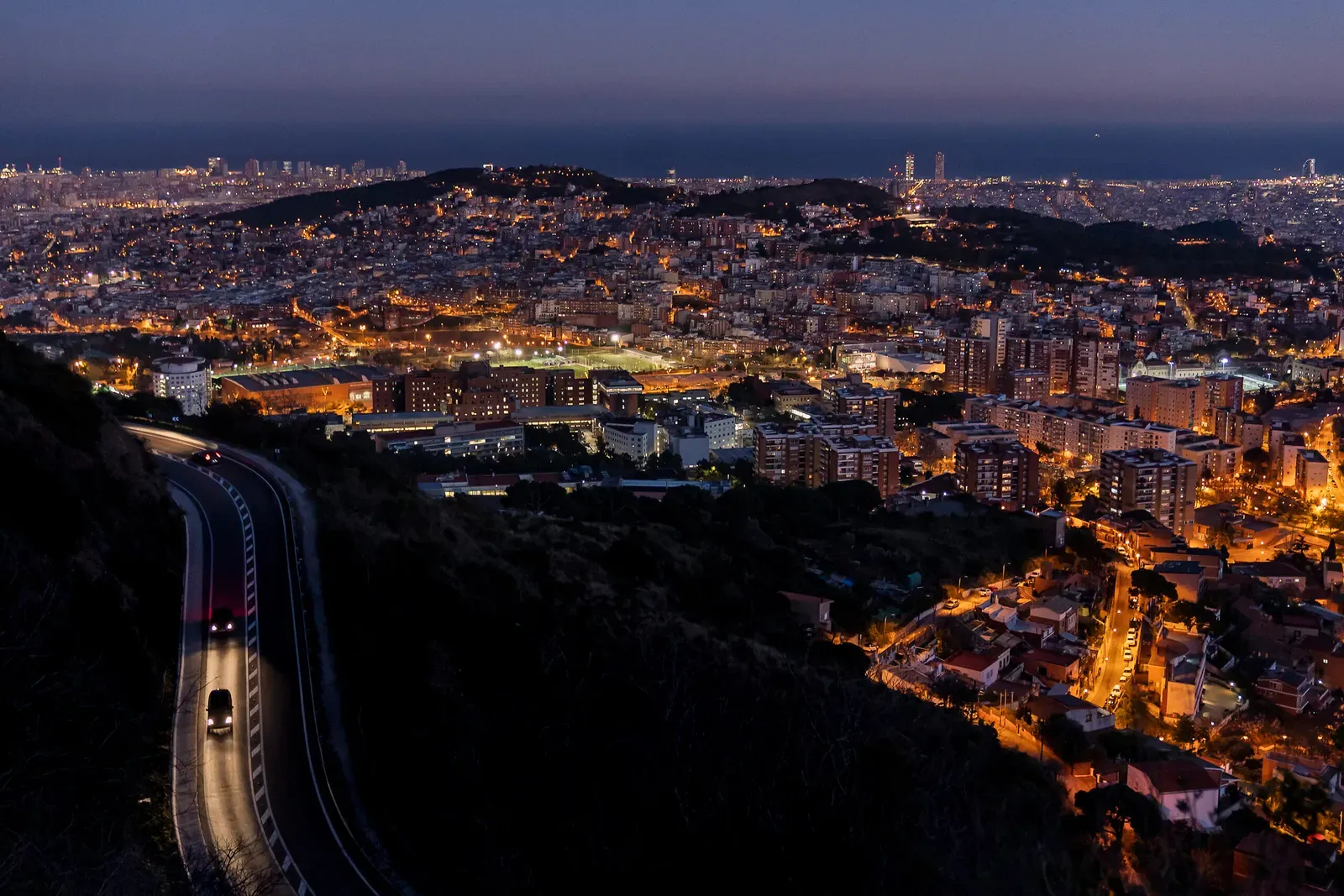 A panoramic night view of Barcelona city with twinkling lights and a road leading into a tunnel, showcasing the urban landscape against a backdrop of hills and a dusky sky.