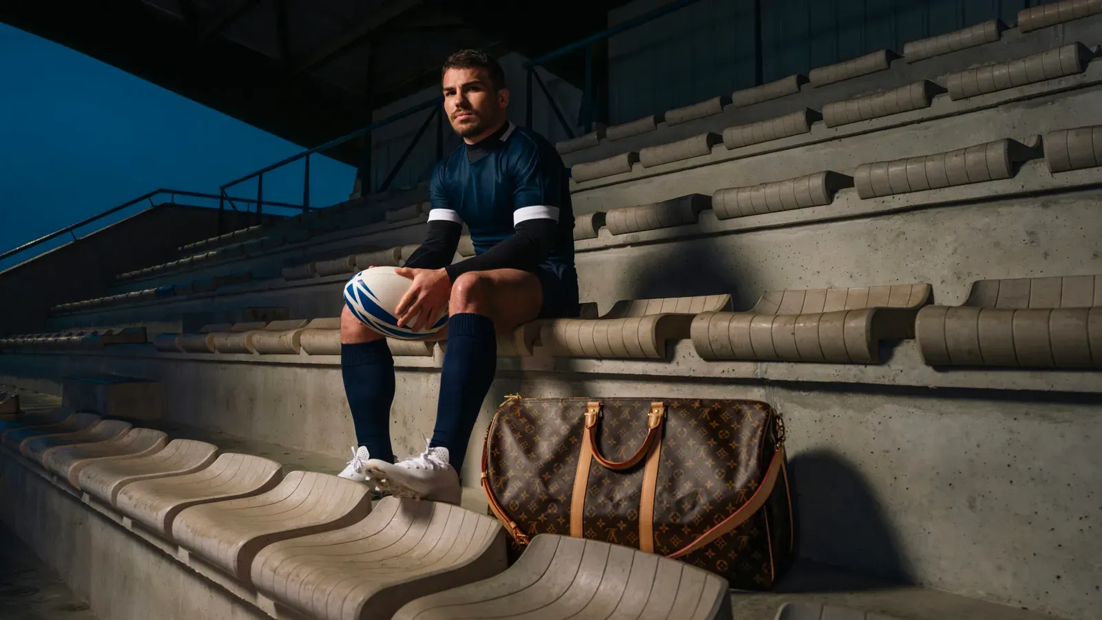 House Ambassador Antoine Dupont, wearing a navy rugby kit, sits on stadium steps with a Louis Vuitton bag beside him and a rugby ball in hand, reflecting the union of sportsmanship and luxury.