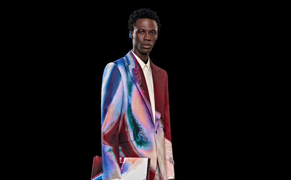 A model presents an Alexander McQueen AW23 avant-garde blazer with a holographic sheen, reflecting a spectrum of vivid colors, paired with a deep red garment underneath, capturing the innovative edge of high fashion.
