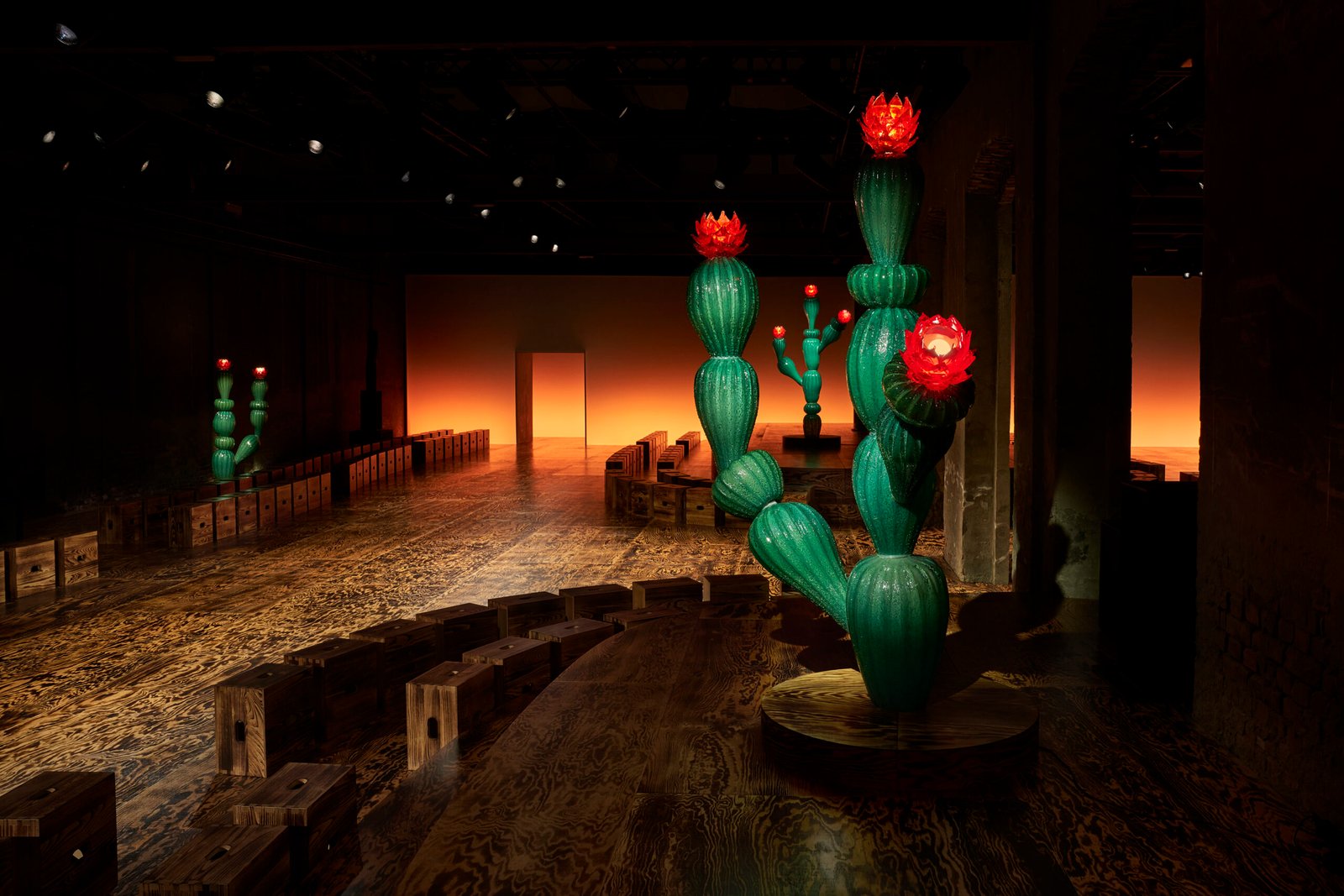 An atmospheric view of Bottega Veneta's Fall/Winter 2024 fashion show venue, featuring artistic cactus sculptures with bright red blooms. The room is dimly lit, highlighting the sculptures and the warm, fiery backdrop at the end of the runway.