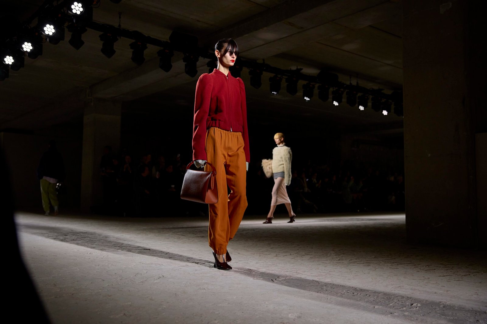 A model parades a Dries Van Noten outfit with a rich red sweater and caramel trousers, accessorized with a brown handbag, on the runway.