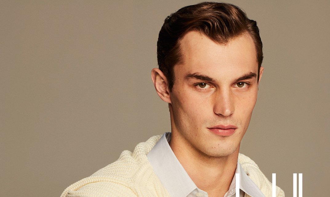 Kit Butler from the dunhill Spring/Summer 2024 Collection poses with effortless elegance. He is wearing a crisp, white button-up shirt with a cream cashmere sweater casually draped over his shoulders. The look is completed with a dark brown leather belt.