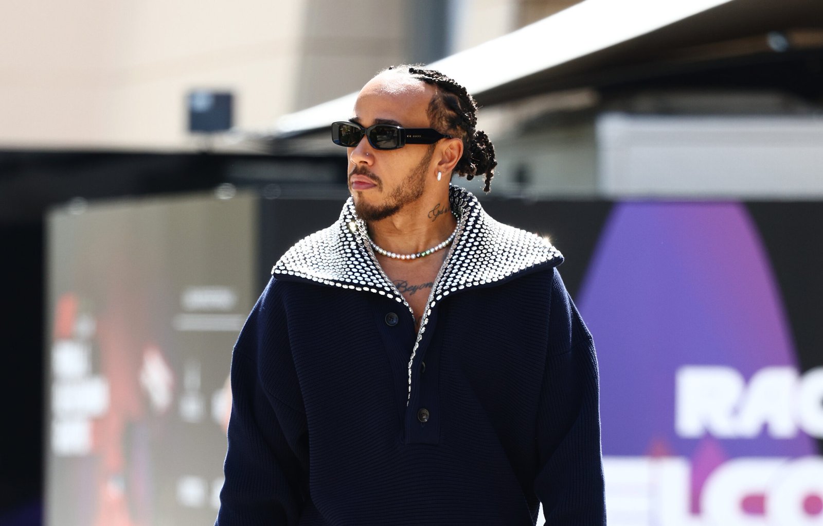Lewis Hamilton strides confidently wearing a navy Gucci sweater with English rib knit texture, dark blue tailored denim pants, Gucci rubber sole mocassins, and stylish Gucci sunglasses at the Bahrain Grand Prix paddock.