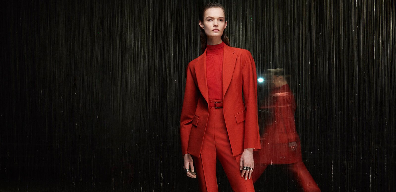 A model dons a tailored red turtleneck ensemble with a structured jacket and belted waist from the Hermès Fall 2024 collection, contrasting with a blurred figure in motion in the background, all set against a rain-like vertical backdrop.