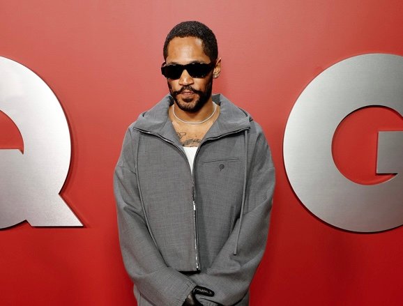 Kaytranada stands confidently on the GQ "Men of the Year" emblem, wearing a Ferragamo fog-grey wool zip-up jacket and trousers with stylish accessories, exuding a vibe that is both laid-back and fashion-forward.