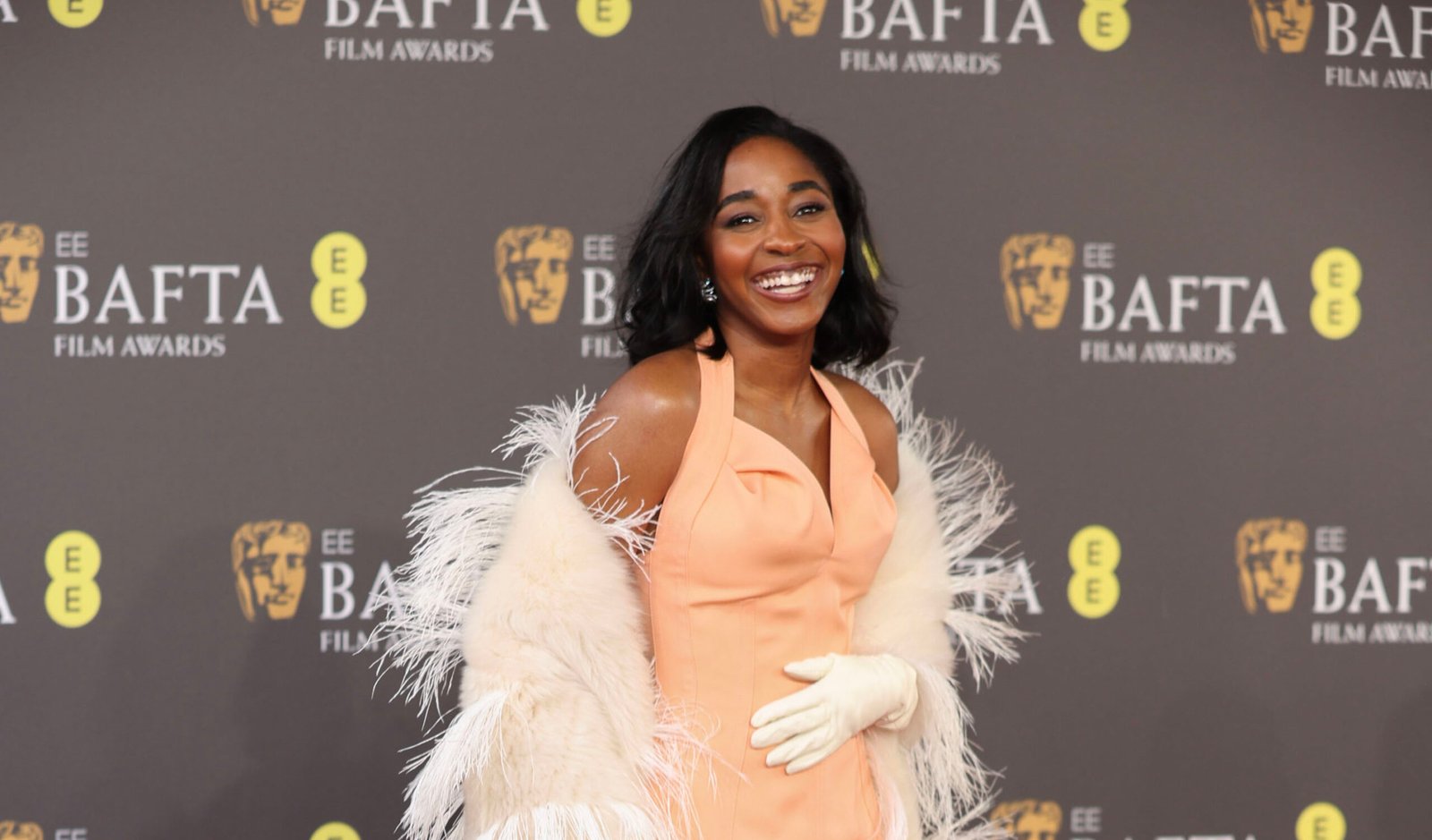Ayo Edebiri stands on the red carpet at the 2024 BAFTA Film Awards, radiant in a bespoke Bottega Veneta peach gown with a luxuriously plumed shawl.
