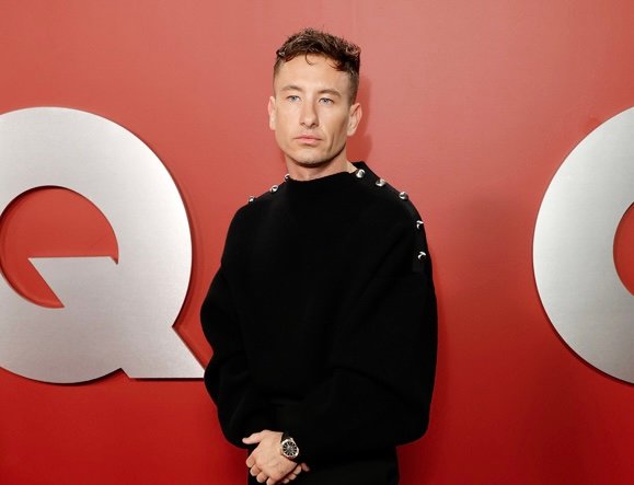 Barry Keoghan stands on the GQ "Men of the Year" emblem, dressed in a black Ferragamo sweater with jewel embellishments and tailored trousers, exuding a blend of modern sophistication and timeless style.