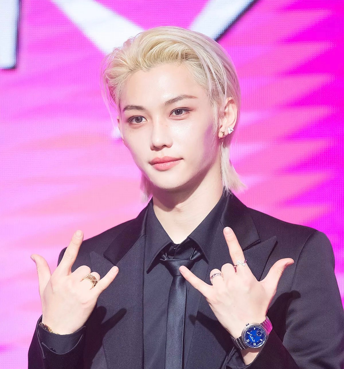 Felix of Stray Kids at a press conference, dressed in a Louis Vuitton black tuxedo and accessorized with fine jewelry, against a pink backdrop.