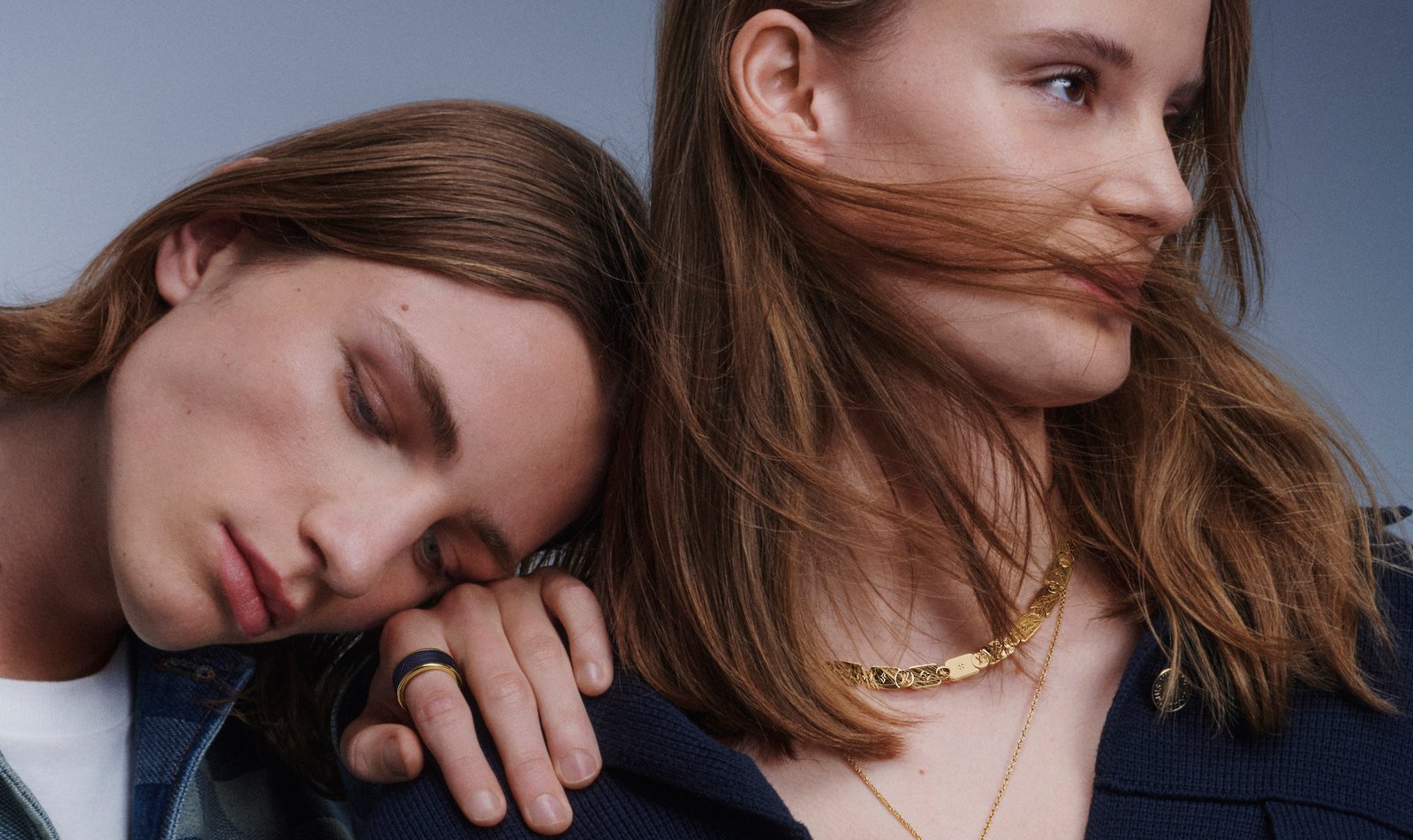 Two individuals close together, one resting their head on the other's shoulder, showcasing Louis Vuitton fine jewelry including a gold necklace and rings.