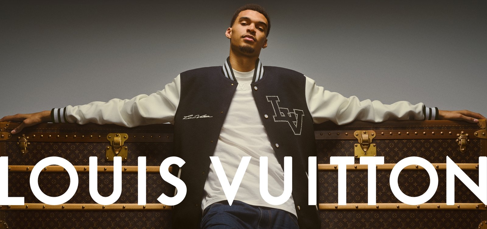 A poised Victor Wembanyama stands confidently with arms extended, resting on a classic Louis Vuitton trunk. He wears a varsity jacket with LV insignia, embodying the fusion of high fashion and athletic elegance.