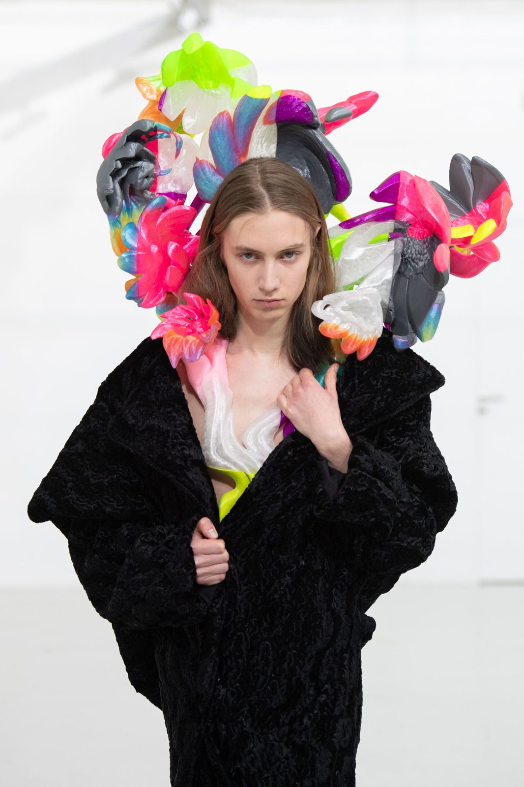 A model from MAITREPIERRE's Autumn/Winter 2024-2025 collection wears an extravagant headpiece with vibrant, multicolored floral shapes, complemented by a textured black coat.