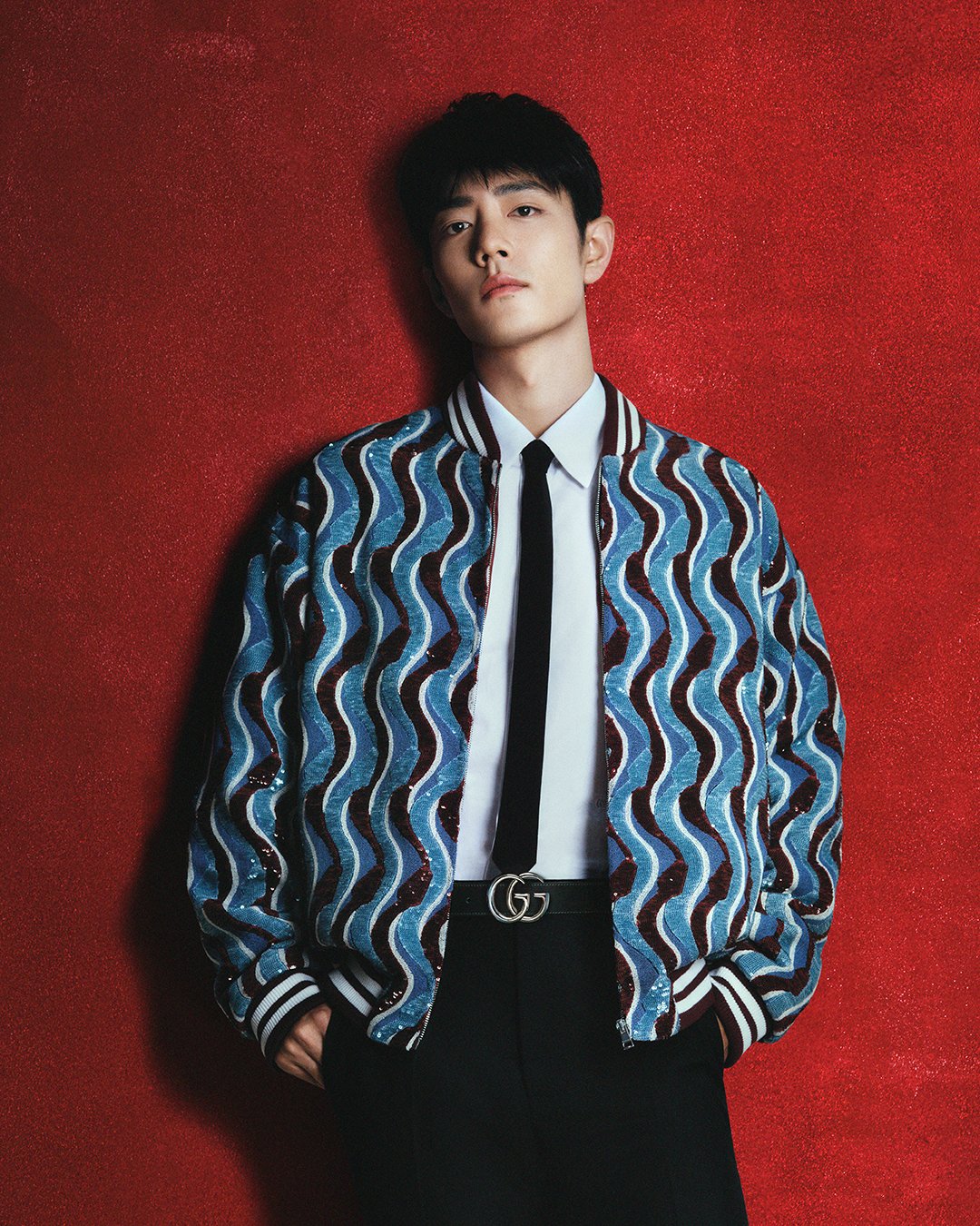 Xiao Zhan wearing a sequined bomber jacket with undulating blue waves, styled with a white dress shirt, black tie, and GG Marmont belt. (Image: Courtesy of Gucci)