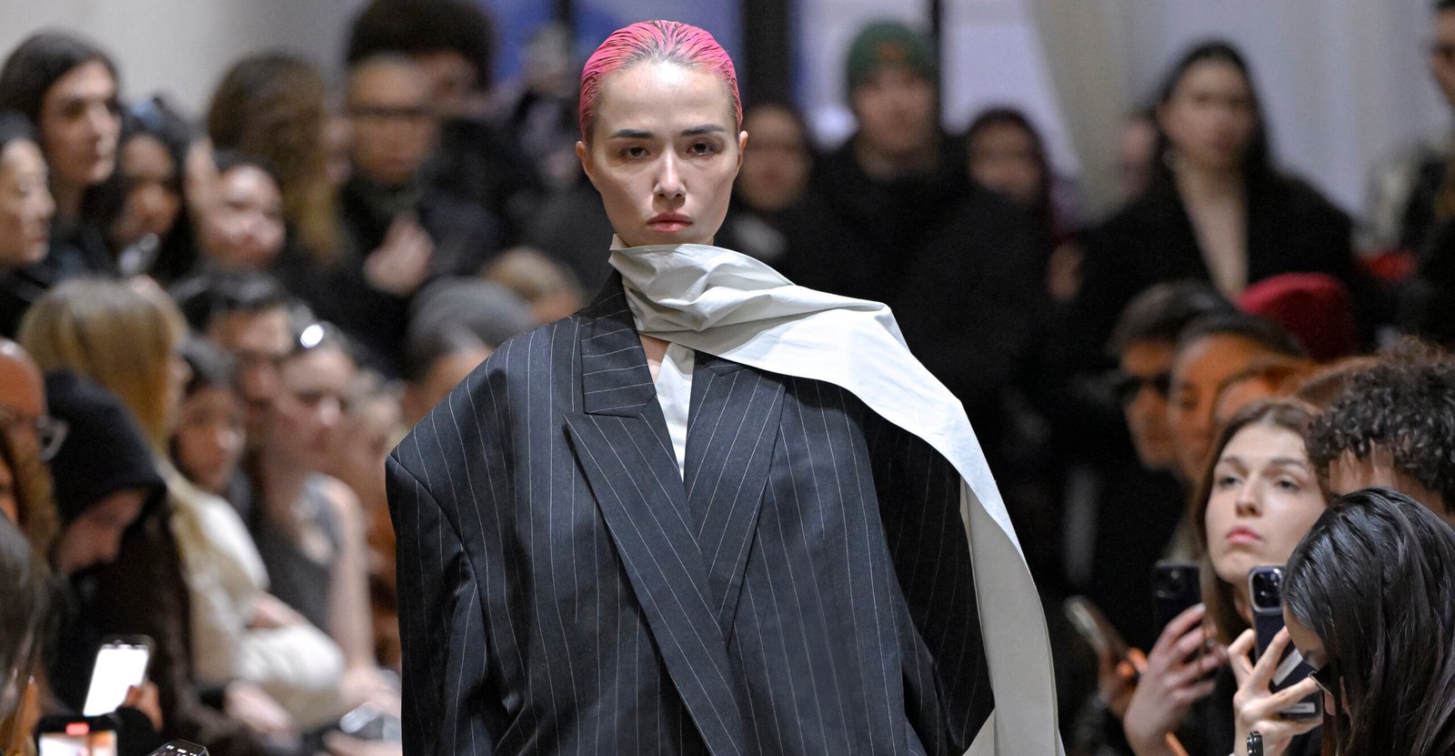 A model exhibits a bold pinstripe blazer with a draped cream scarf from Vautrait's Fall/Winter 2024 collection on the runway, complemented by a stark pink hair color, encapsulating the brand's avant-garde approach to sustainable fashion.