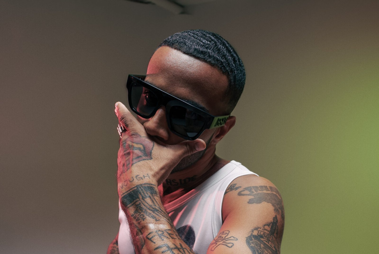 Vic Mensa poses with a hand over his mouth, wearing sunglasses and a white tank top, his tattoos on full display, creating a powerful visual statement.