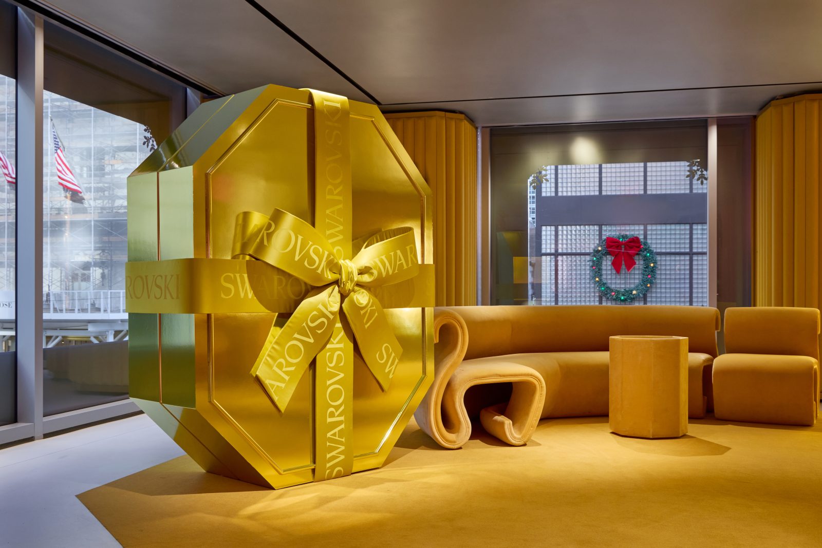 A golden gift box with the signature Swarovski ribbon takes center stage in a modern lounge, while a holiday wreath connects the interior to the city beyond. (Photo: Courtesy of Swarovski)
