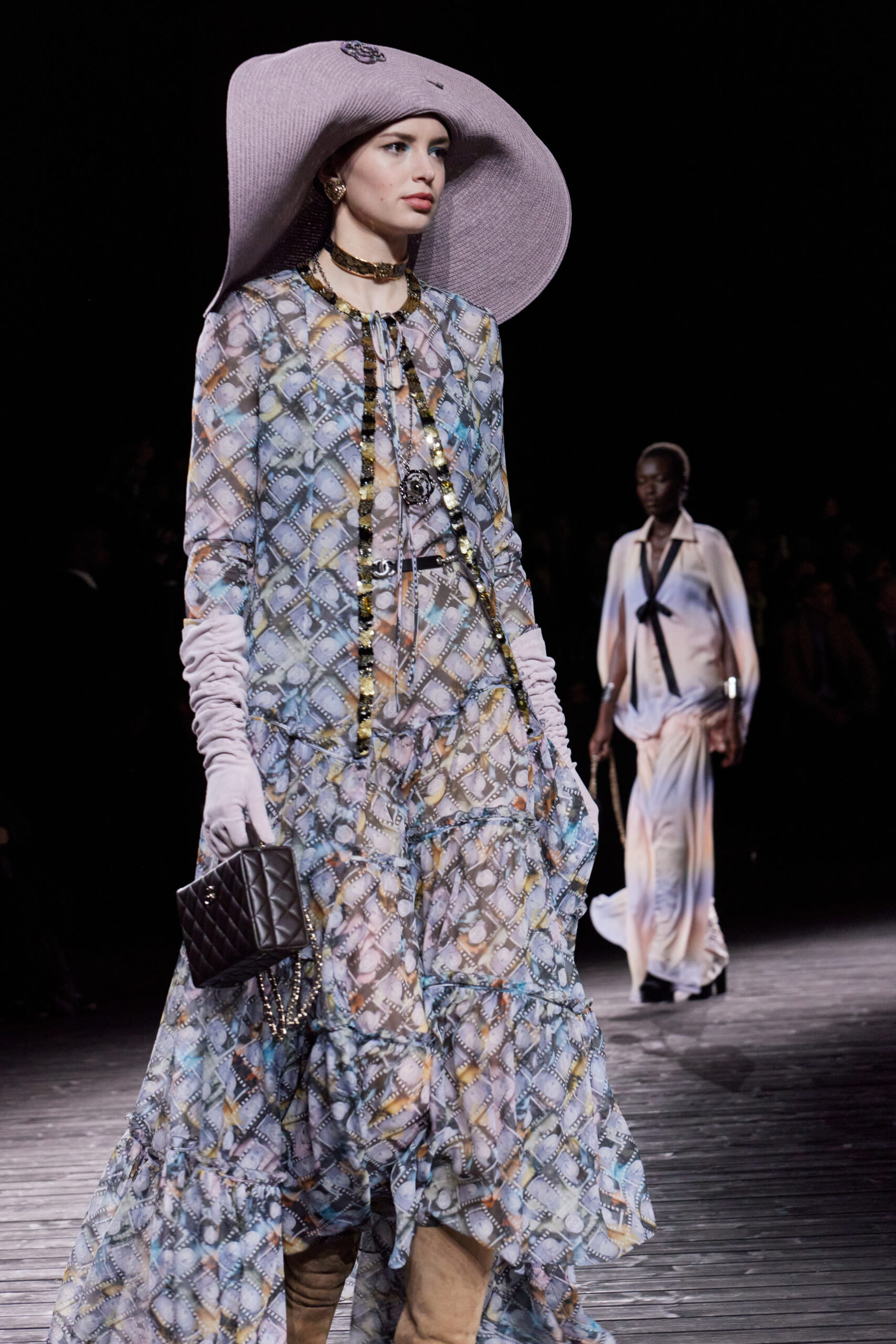 Virginie Viard’s indelible Fall/Winter 2024 collection transported CHANEL back to the brands origins on the promenades of Deauville. A model walks the runway at the CHANEL Fall/Winter 2024/25 show in a pastel patterned dress with film motif, paired with a wide-brimmed hat, accessorized with layered necklaces and a quilted handbag.