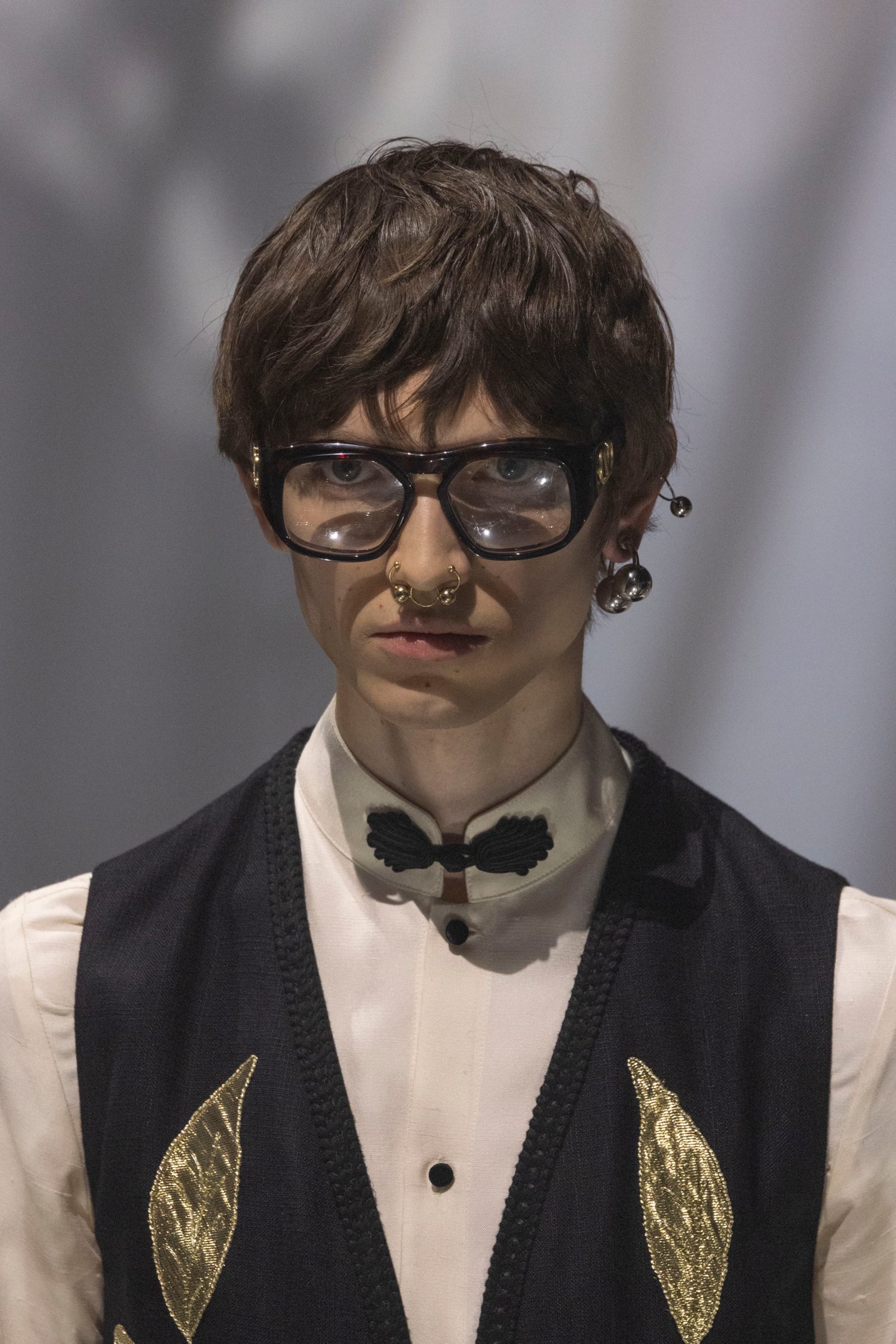 Close-up of model in chunky black Valentino optical frames with gold VLogo detail and dangling charms, wearing black vest with gold embroidered details