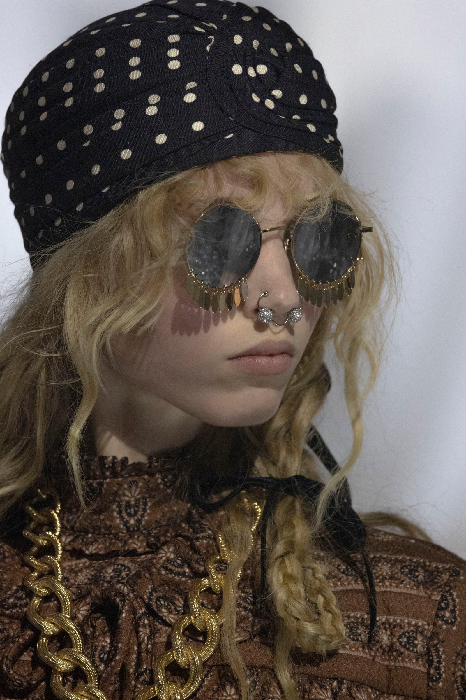 Model wearing Valentino gold-trimmed round sunglasses with decorative fringe detail, polka dot headscarf, and brown lace-detailed top with gold chain accents.