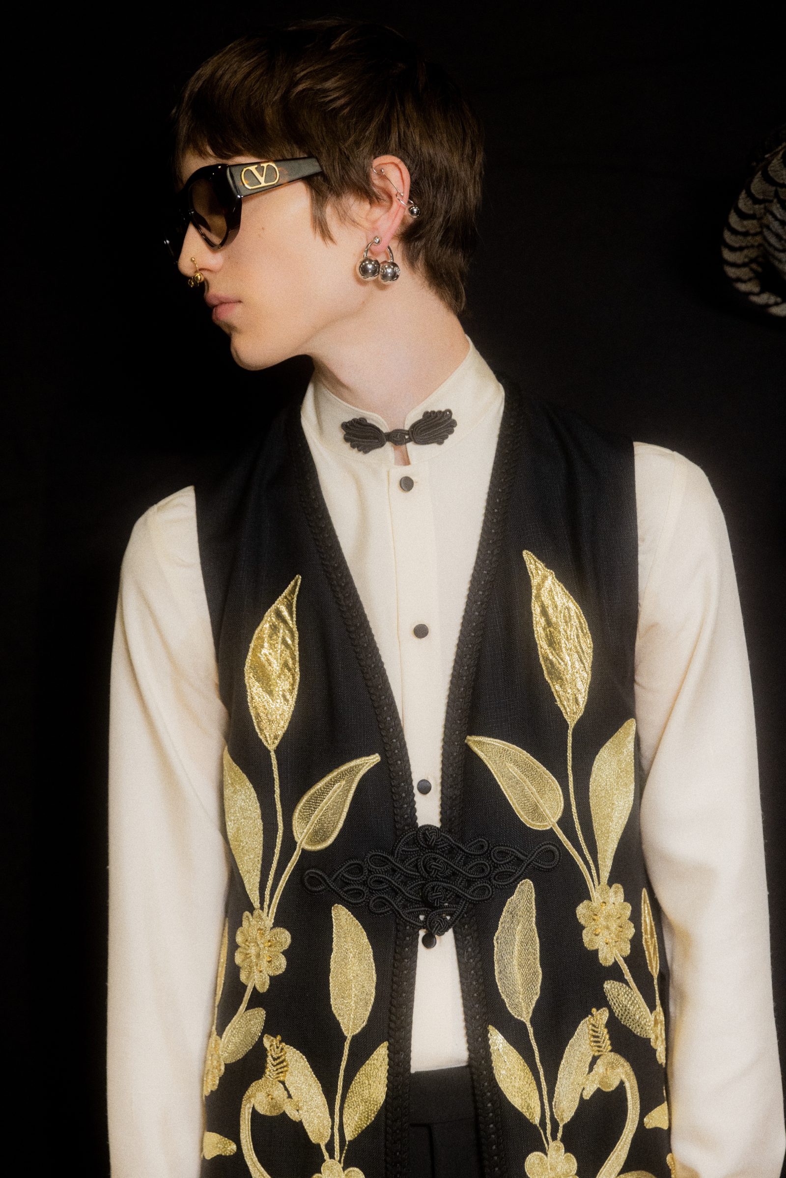 Model in profile wearing black Valentino sunglasses with VLogo detail, black vest with gold floral embroidery, and white shirt with black butterfly bow tie. Dangling silver ball earrings complete the look