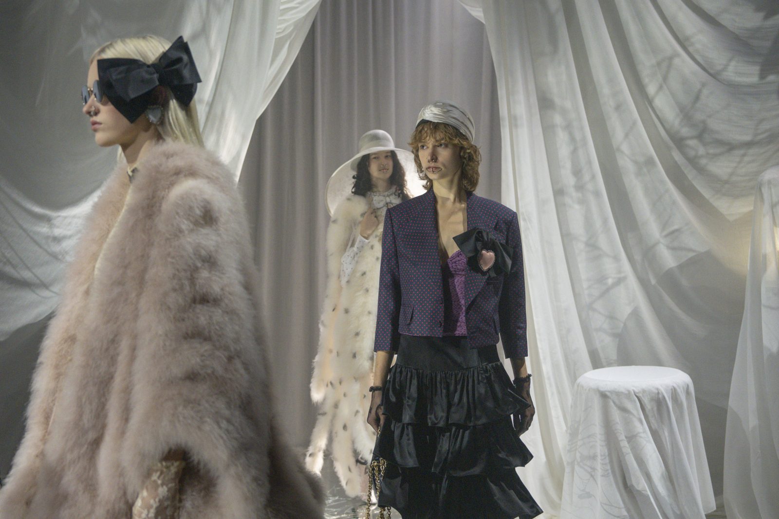 The Valentino Spring-Summer 2025 presentation unfolds against white draping: a cropped blazer with bow embellishment meets cascading black silk ruffles, while a blush plumage coat and spotted organza gown frame the scene.