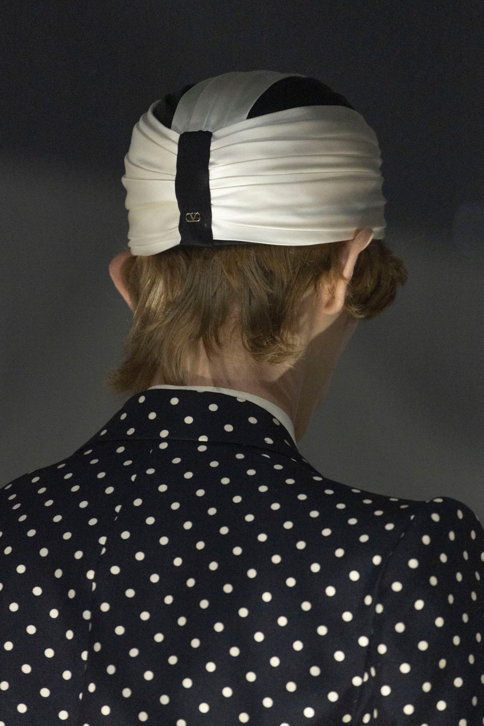 Polka dots complement a pleated satin turban, secured with Valentino's monogrammed band, creating a sophisticated nod to 1920s glamour.