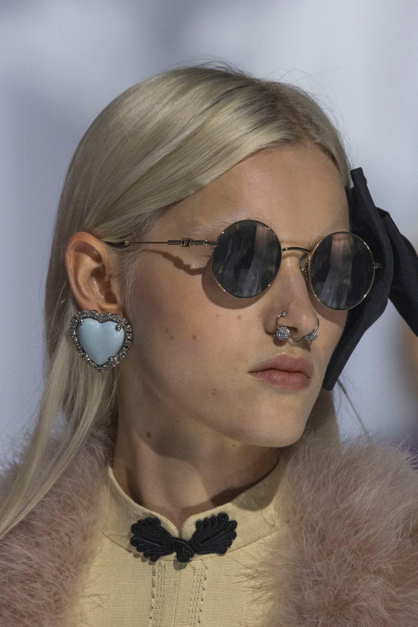 Past meets present: round gold-rimmed sunglasses and a black satin bow, while a crystal-trimmed powder blue heart earring and septum ring echo the collection's romance-meets-rebellion aesthetic.