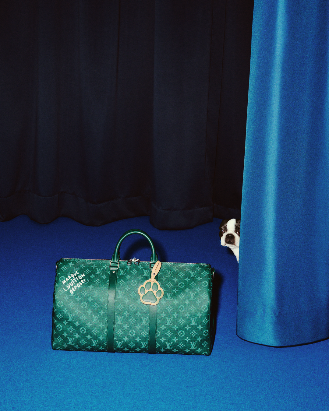 A playful peek-a-boo moment: a Boston Terrier peeks from behind a curtain at Louis Vuitton's green Monogram Keepall adorned with a paw print charm from the Dog LVππππ Pre-Collection. (Photo: Courtesy of Louis Vuitton)