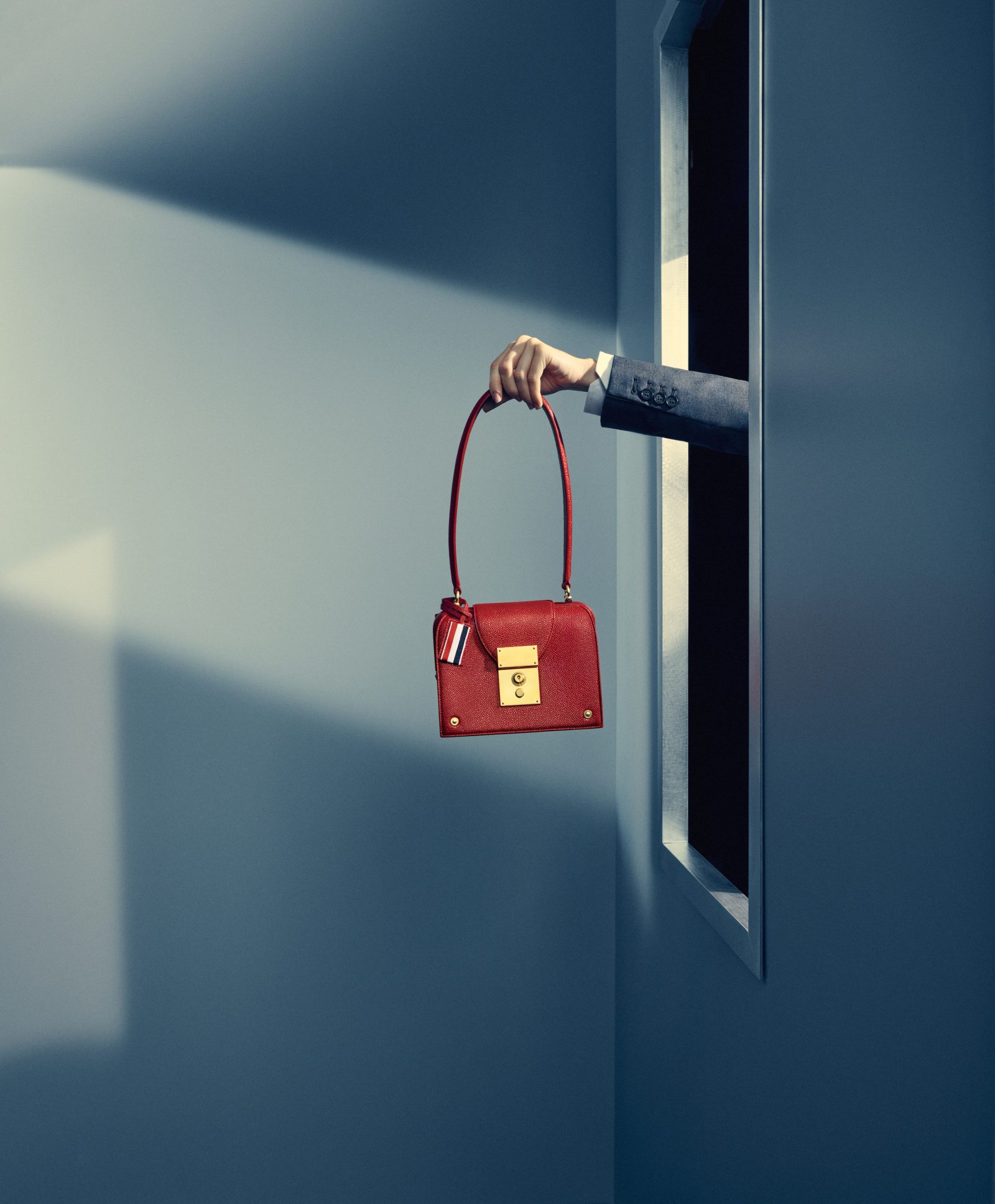 A striking gesture frames the Mrs. Thom Triangle Bag in crimson, its polished hardware catching the light as it floats within Browne’s surrealist elevator scene. (Image: Courtesy of Thom Browne)