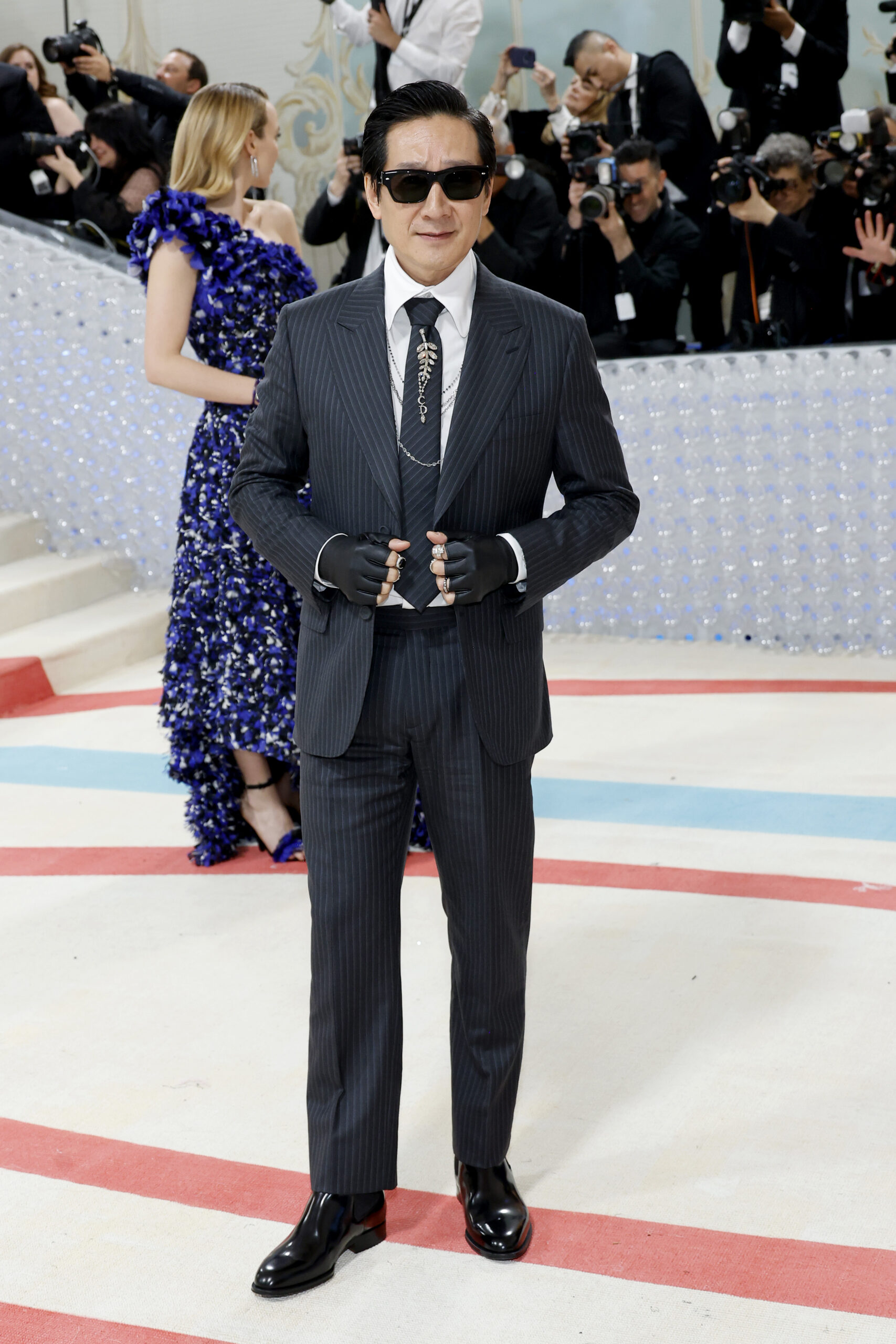 Ke Huy Quan wore a custom made Dior men’s collection black striped wool peak lapel suit, a white cotton wide collar shirt, a black striped silk wide tie, a silver brooch with CD letters, black leather fingerless gloves and black leather boots