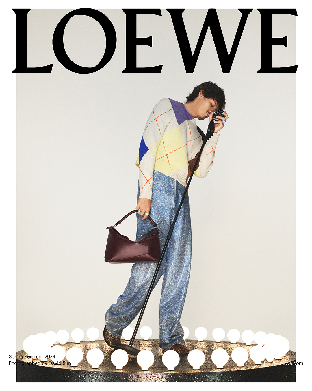Omar Apollo in a vibrant, abstract knit sweater and shimmering denim, poised with a camera and the new Loewe Puzzle bag, encapsulates the artistic spirit on a stage of illuminated spheres. Photographed by David Sims for Loewe.