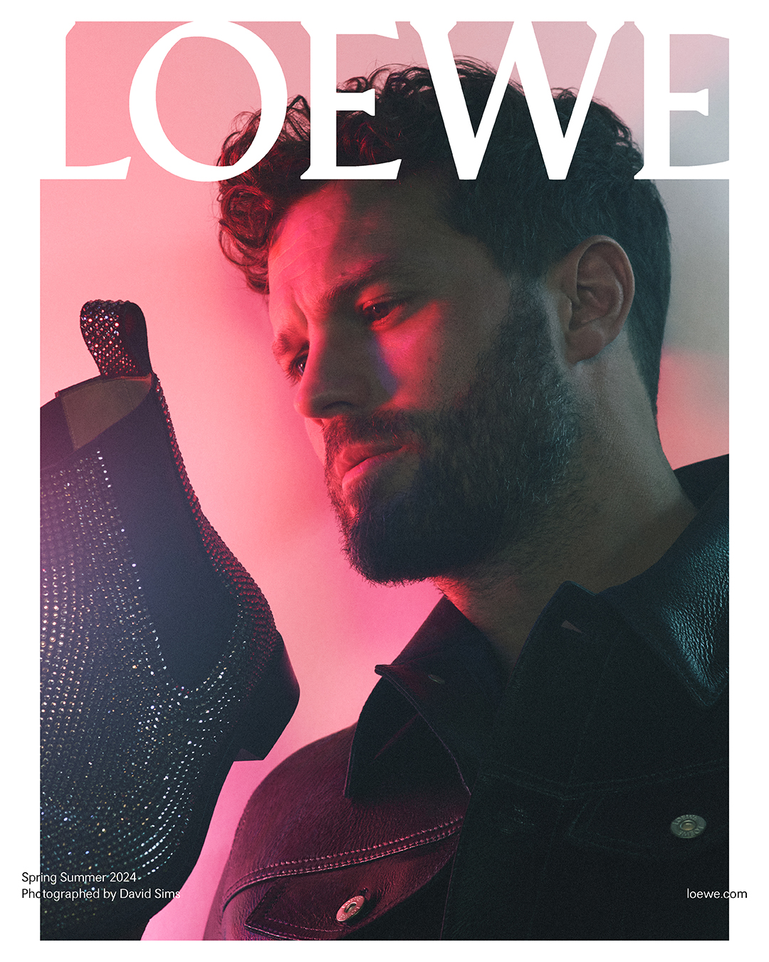 Jamie Dornan, as Loewe's Global Brand Ambassador, wears a textured leather jacket, meditatively holding a sparkling Loewe boot, under a roseate hue. The Spring/Summer 2024 campaign by David Sims captures the essence of luxury.