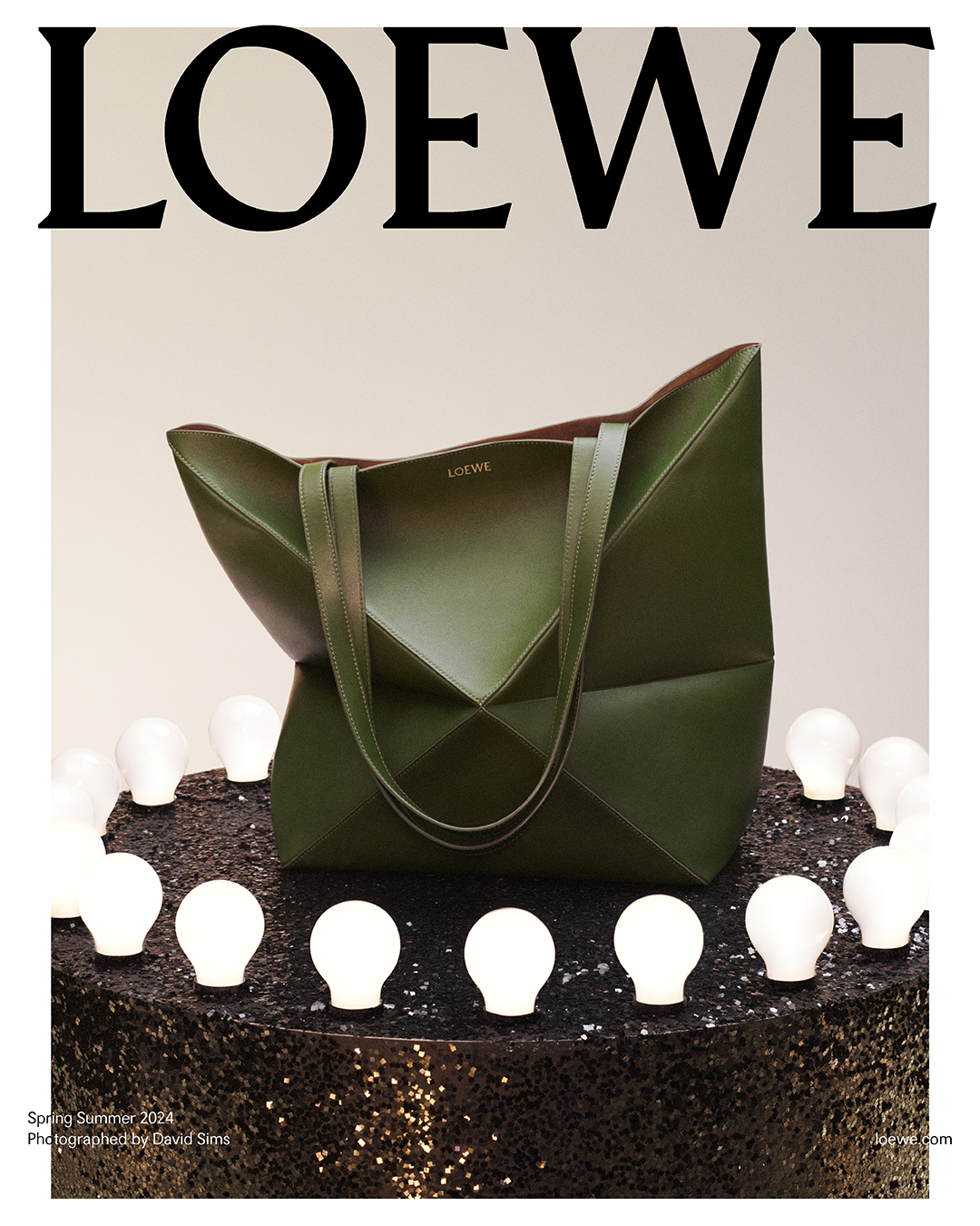 The iconic Loewe Puzzle tote in olive green stands out against a backdrop of glowing orbs, resting on a glittering podium, symbolizing the intersection of innovative design and timeless beauty. Photographed by David Sims for Loewe.