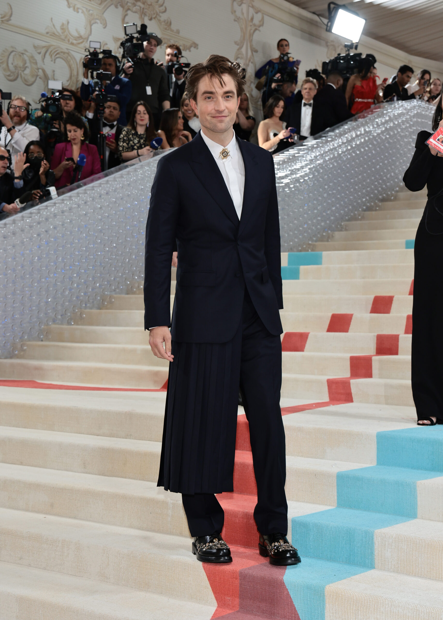 Robert Pattinson wore a custom made Dior Fall 23 men’s collection midnight blue virgin wool and linen peak lapel suit, a midnight blue wool and linen long kilt, a white cotton classic collar shirt, a gold brooch and black leather Carlo derbies