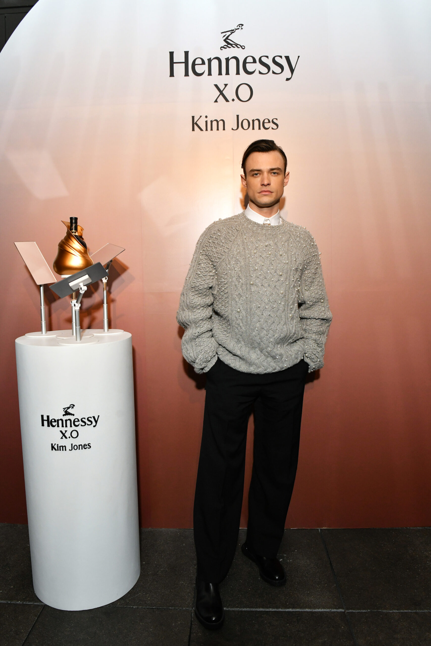 Thomas Doherty wore a Dior men’s 2023 ERL capsule collection grey wool and cashmere crewneck cable knit sweater with pearl dégradé embroidery, a white cotton shirt and black wool pants. Thomas Doherty