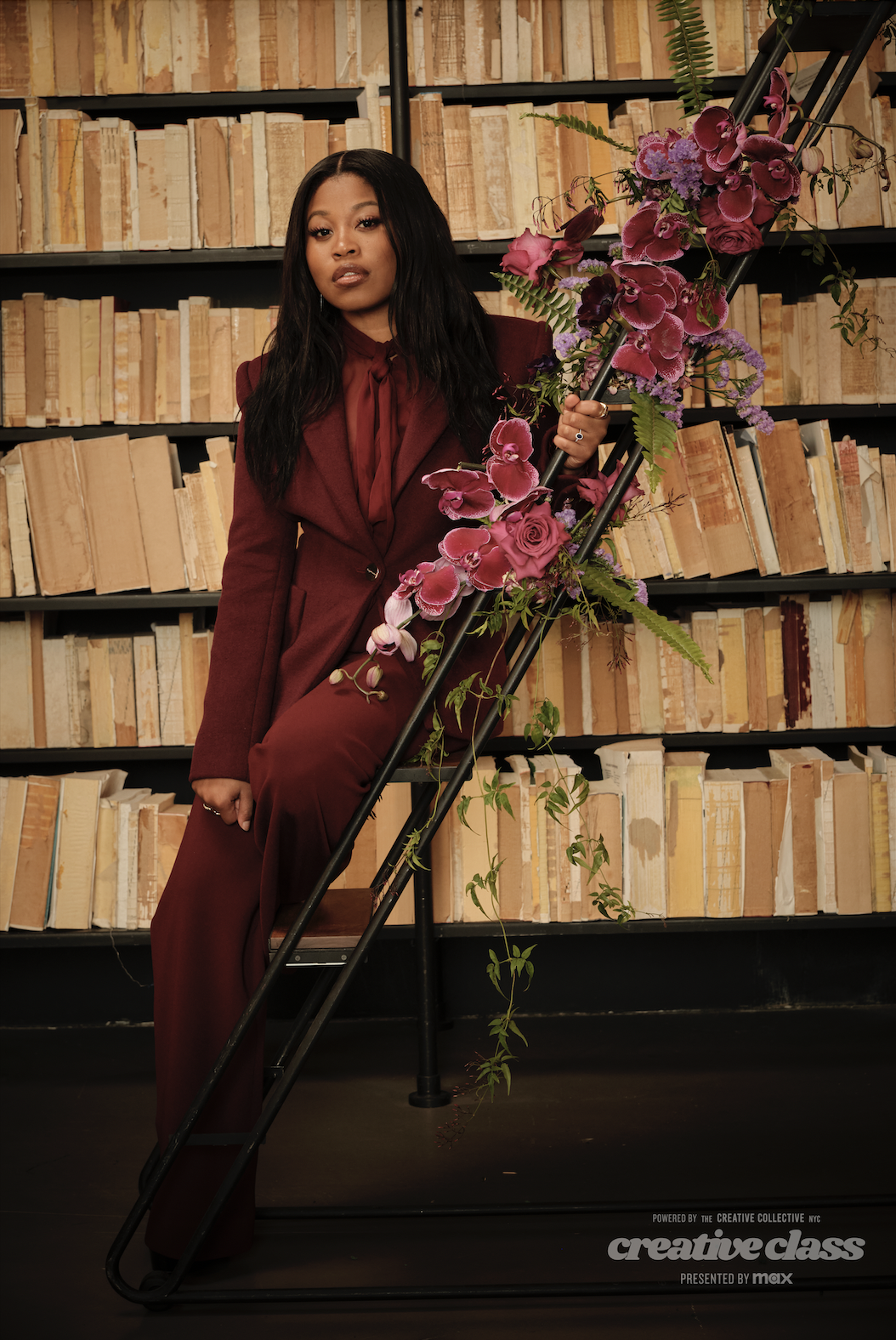 Against a backdrop of timeless literature, Dominique Fishback poses at the Creative Collective NYC event, her Sergio Hudson maroon suit perfectly complementing her poise and rising star status. (Photo Credit: Rashida Zagon)